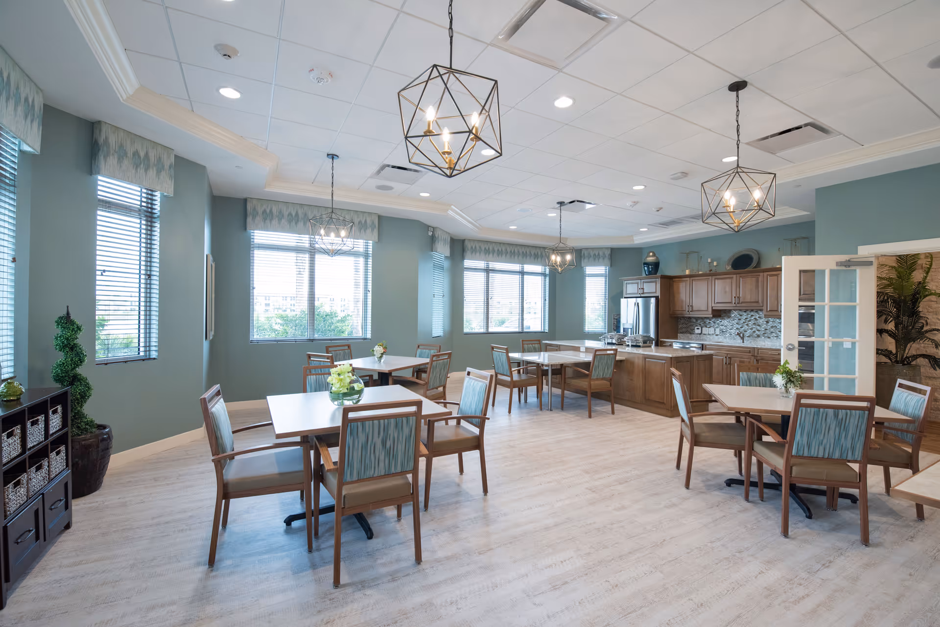 A bright and spacious dining room with several square tables and wooden chairs with blue patterned cushions. The room features large windows with blinds, light blue walls, and three geometric pendant lights hanging from the ceiling. In the background, there is a kitchen area with wooden cabinets, a stainless steel refrigerator, and a tiled backsplash. Decorative plants and small floral arrangements are placed on the tables and around the room.