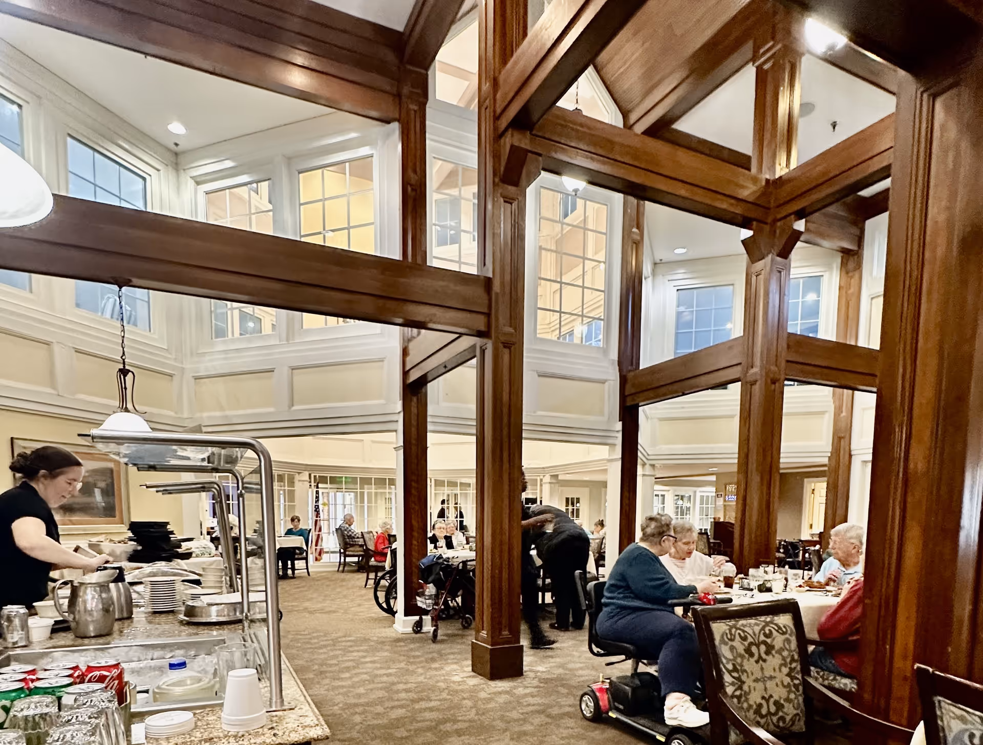 A spacious dining room in a senior living facility with high ceilings and large windows. Several elderly residents are seated at tables, eating and socializing. A staff member is seen serving or clearing dishes near a counter with beverages and plates. The room features wooden beams and columns, carpeted floors, and comfortable chairs.