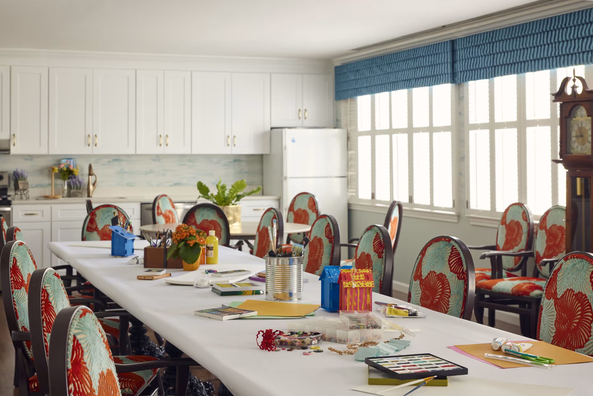 A bright room with a long table covered in a white tablecloth, surrounded by chairs with floral upholstery. The table has various craft supplies, including paint, brushes, papers, and small decorative items. The room has large windows with blue valances, white cabinetry, a refrigerator, and a tall grandfather clock in the corner.