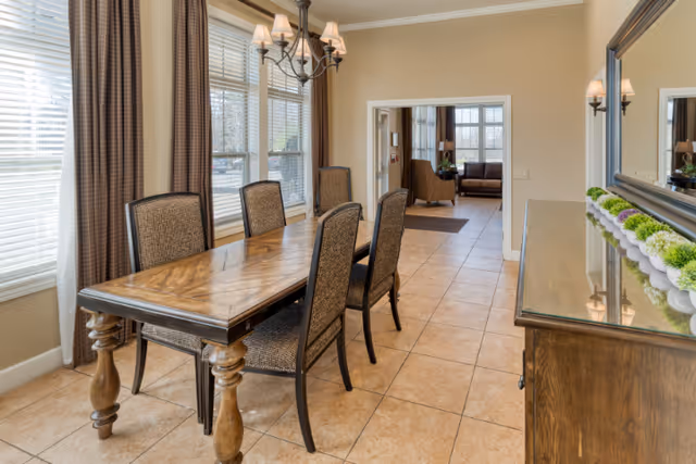A dining room with a wooden table and six upholstered chairs. The room has large windows with brown curtains, beige walls, and tiled flooring. A chandelier hangs above the table. In the background, there is an open doorway leading to a living room area with a sofa and armchairs.