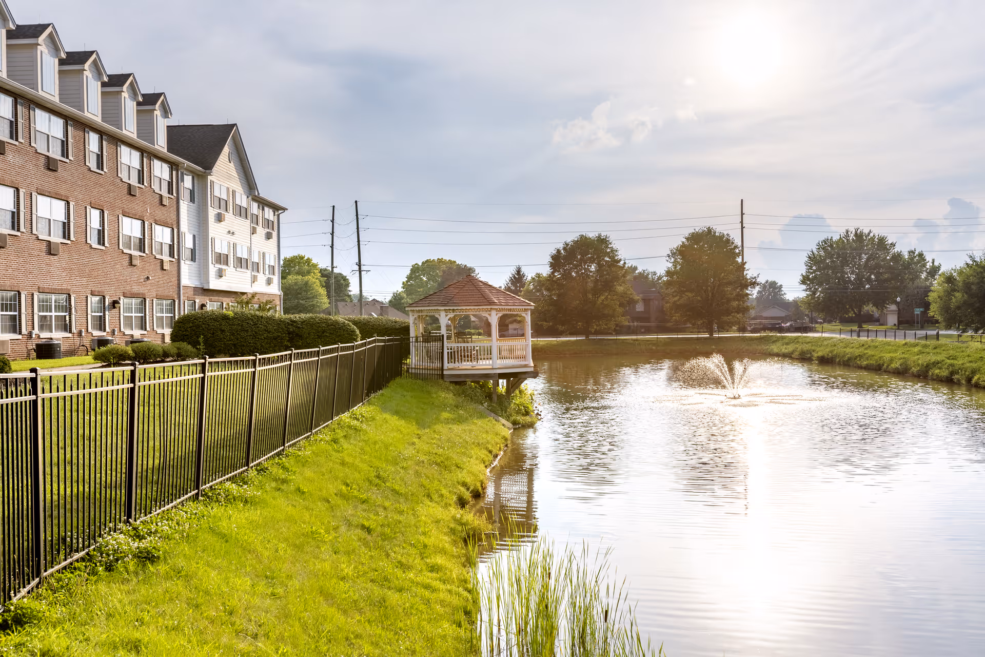 View of a senior living facility building next to a pond with a water fountain. There is a black metal fence along the grassy bank of the pond, and a white gazebo is situated over the water. Trees and a partly cloudy sky are visible in the background.
