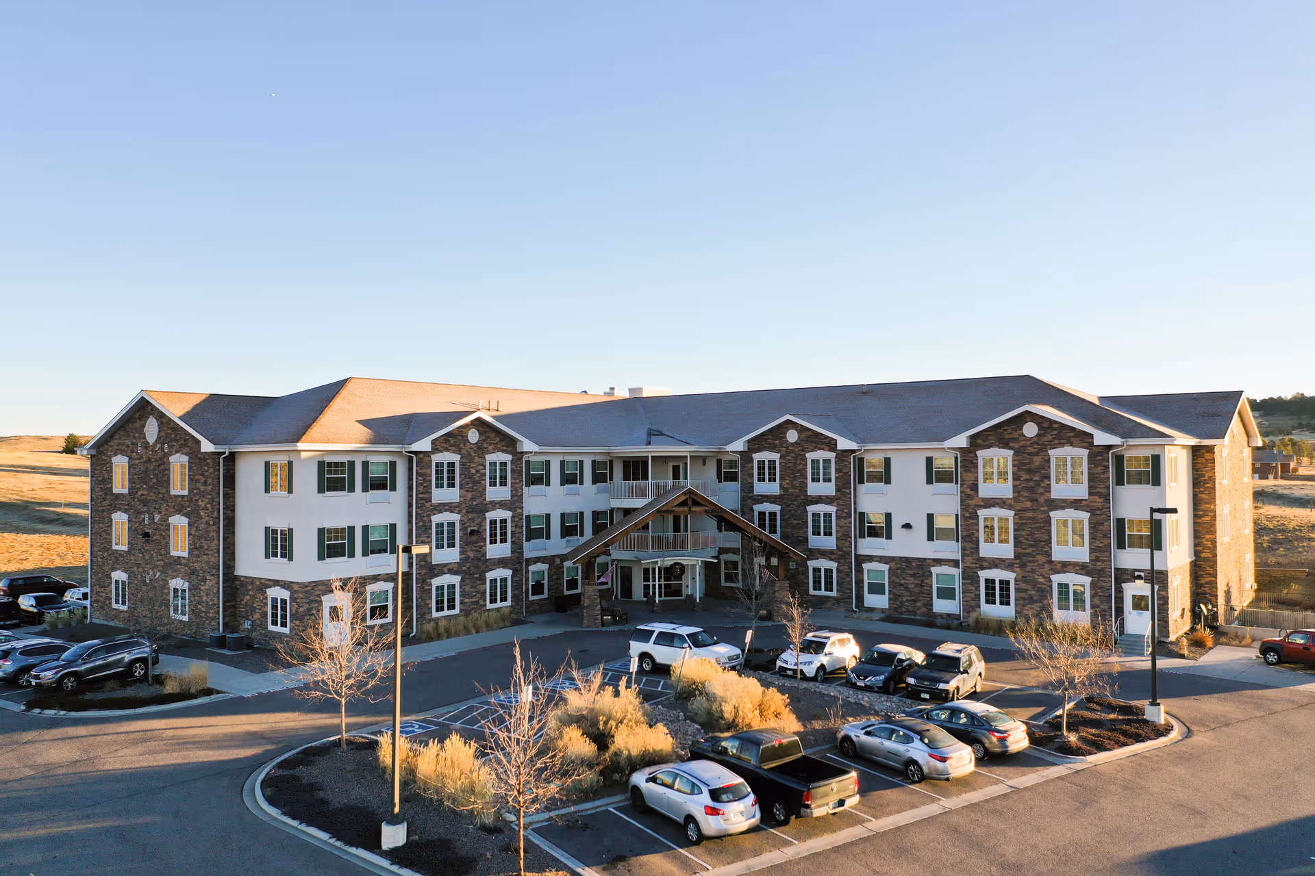 Exterior view of a three-story senior living facility building with stone and white facade, multiple windows, a covered entrance, and a parking lot with several cars parked in front. The surrounding area is open with dry grass and clear sky.