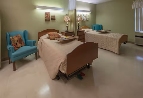 Shared patient bedroom with two hospital-style beds, bedside table and lamps, and upholstered chairs against pale green walls.