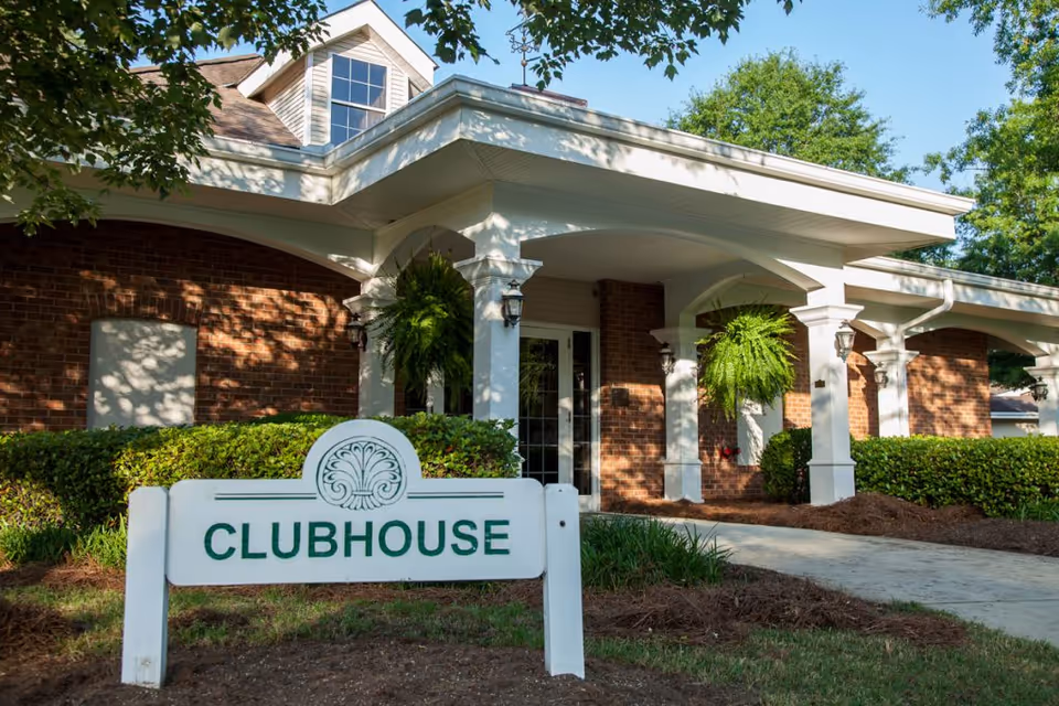 Exterior view of a clubhouse building with brick walls, white columns, and a covered entrance. There are hanging green plants and a white sign in front that reads 'CLUBHOUSE'. The area is surrounded by green bushes and trees under a clear blue sky.
