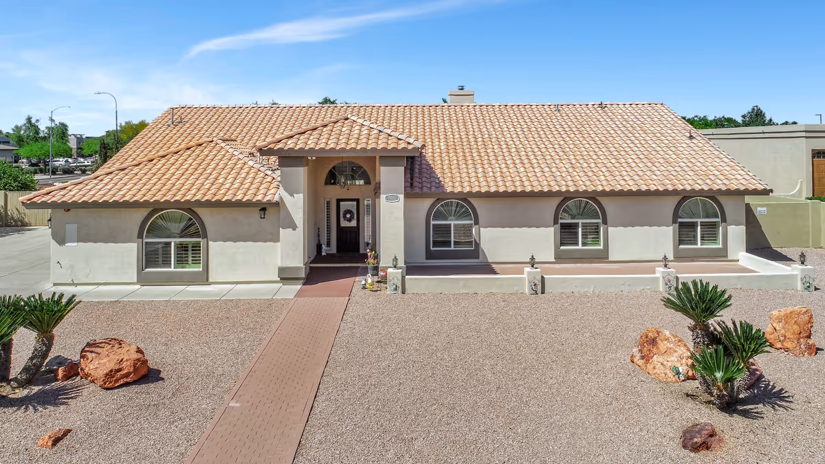 Front exterior view of a single-story building with a tiled roof, arched windows, and a central entrance with a covered porch. The surrounding yard is landscaped with gravel, rocks, and small palm plants under a clear blue sky.