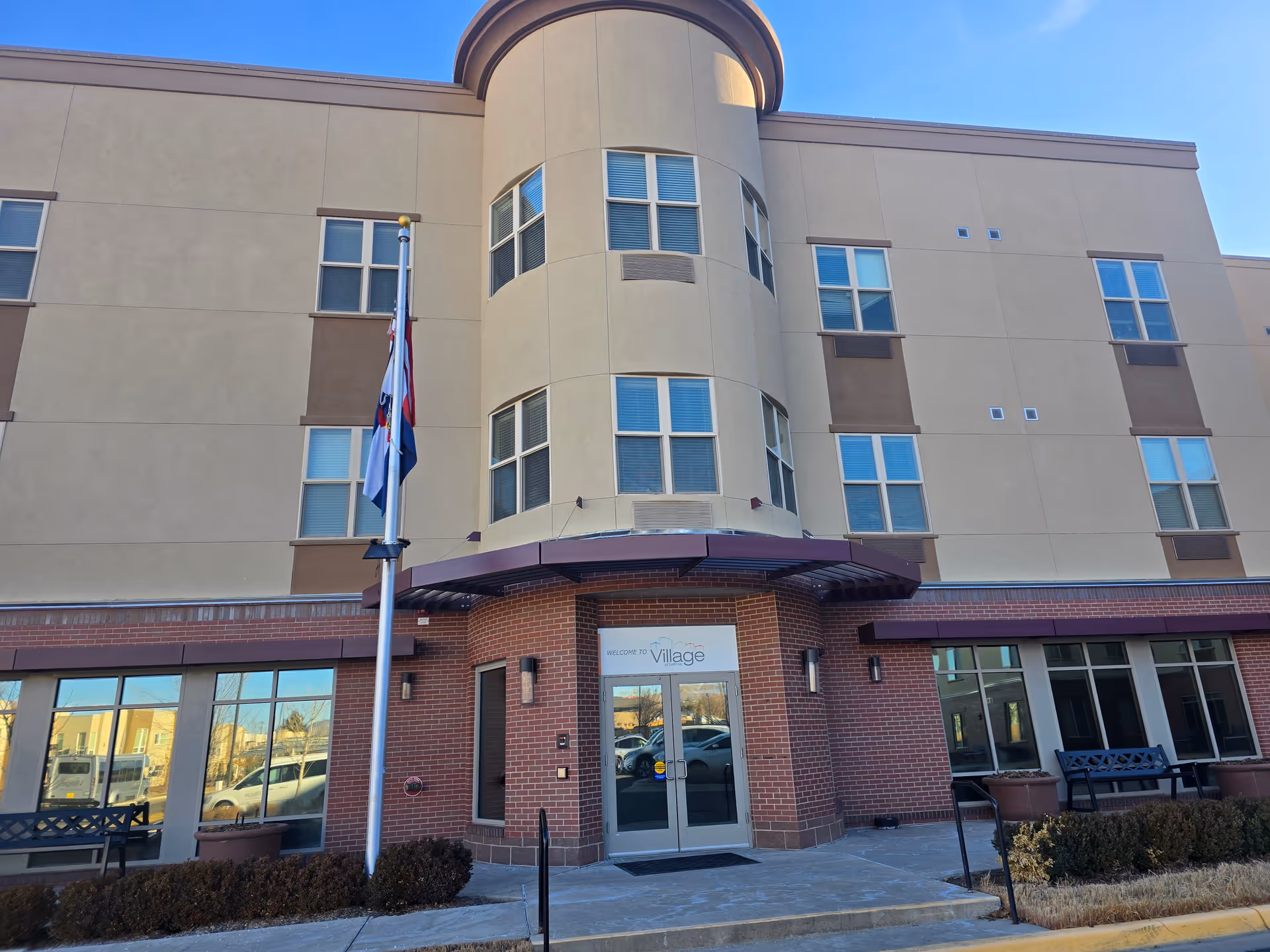 Front entrance of a multi-story senior living building with a flagpole, benches, and glass doors.