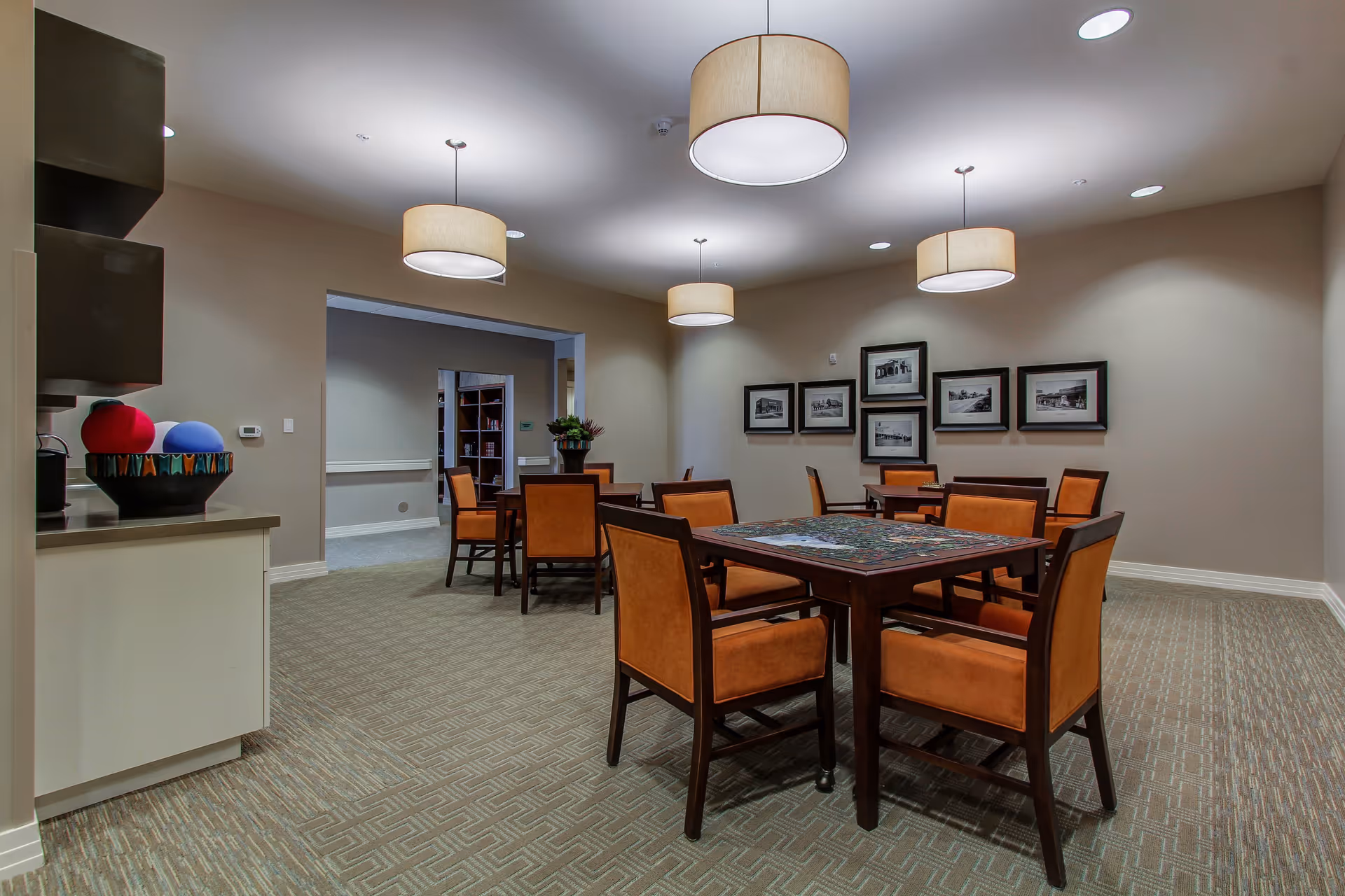 A well-lit room with several wooden tables and orange cushioned chairs arranged around them. The tables have puzzles on them. The walls are beige with framed black and white photographs. Four large round ceiling lights hang from the ceiling. There is a counter with decorative bowls on the left side and an open doorway leading to another room in the background.