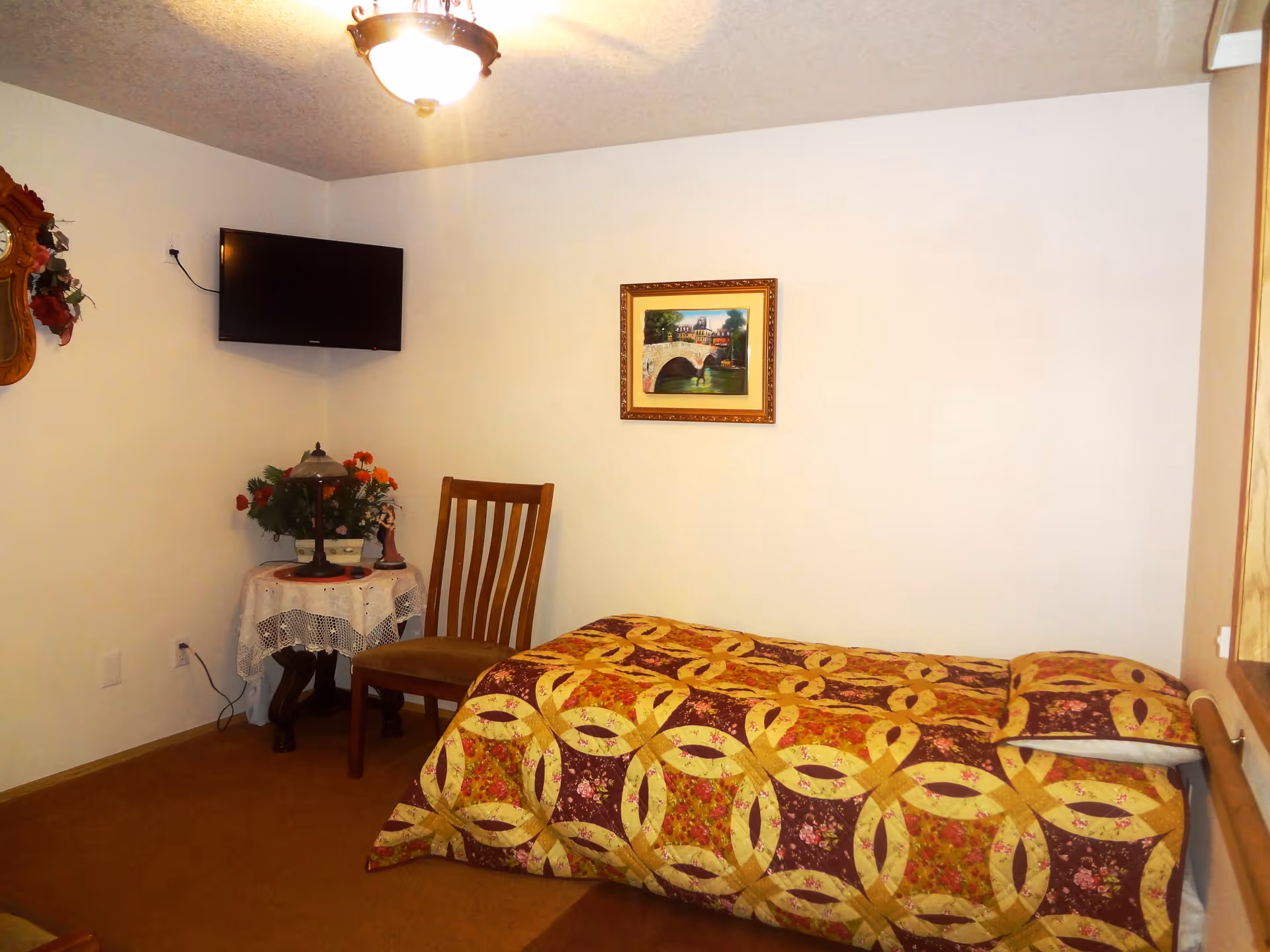 A small bedroom with a single bed covered in a patterned quilt with yellow and maroon colors. There is a wooden chair next to a round table with a lace tablecloth, a lamp, and a flower arrangement. A flat-screen TV is mounted on the wall above the table. A framed painting of a bridge and buildings hangs on the wall above the bed. The room has beige carpet and white walls.