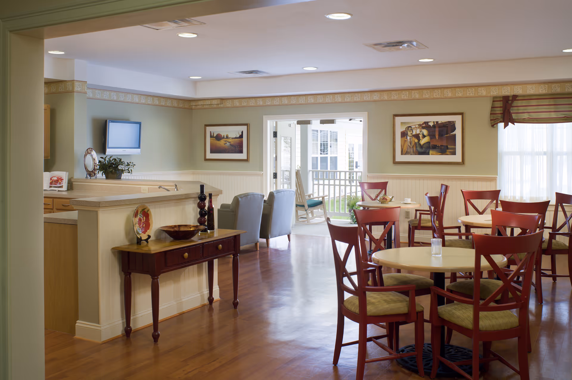 A bright and inviting dining area in a senior living facility with several round tables and red wooden chairs. The room has light green walls with white wainscoting and framed artwork. There is a small wooden console table with decorative items, and a partial view of a kitchen counter with a mounted TV. In the background, there is a seating area with two armchairs and a view of an outdoor porch with rocking chairs.