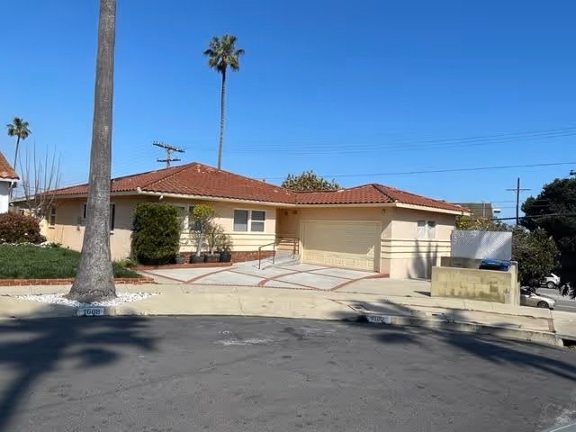 Single-story residential building with a red tile roof and beige exterior walls, situated on a curved street with a palm tree in the front yard and a driveway leading to a garage.