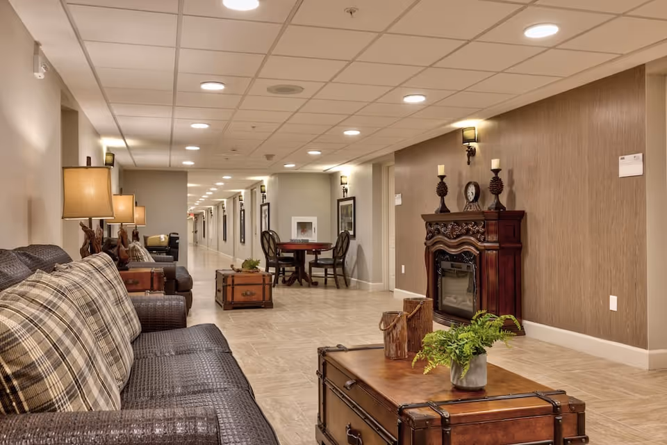 A long hallway in a senior living facility with a seating area on the left featuring a dark wicker sofa with plaid cushions and matching armchairs. There are wooden trunks used as coffee tables, a small round table with chairs further down the hall, and a decorative wooden fireplace with candles and a clock on the right wall. The hallway is well-lit with ceiling lights and wall sconces.
