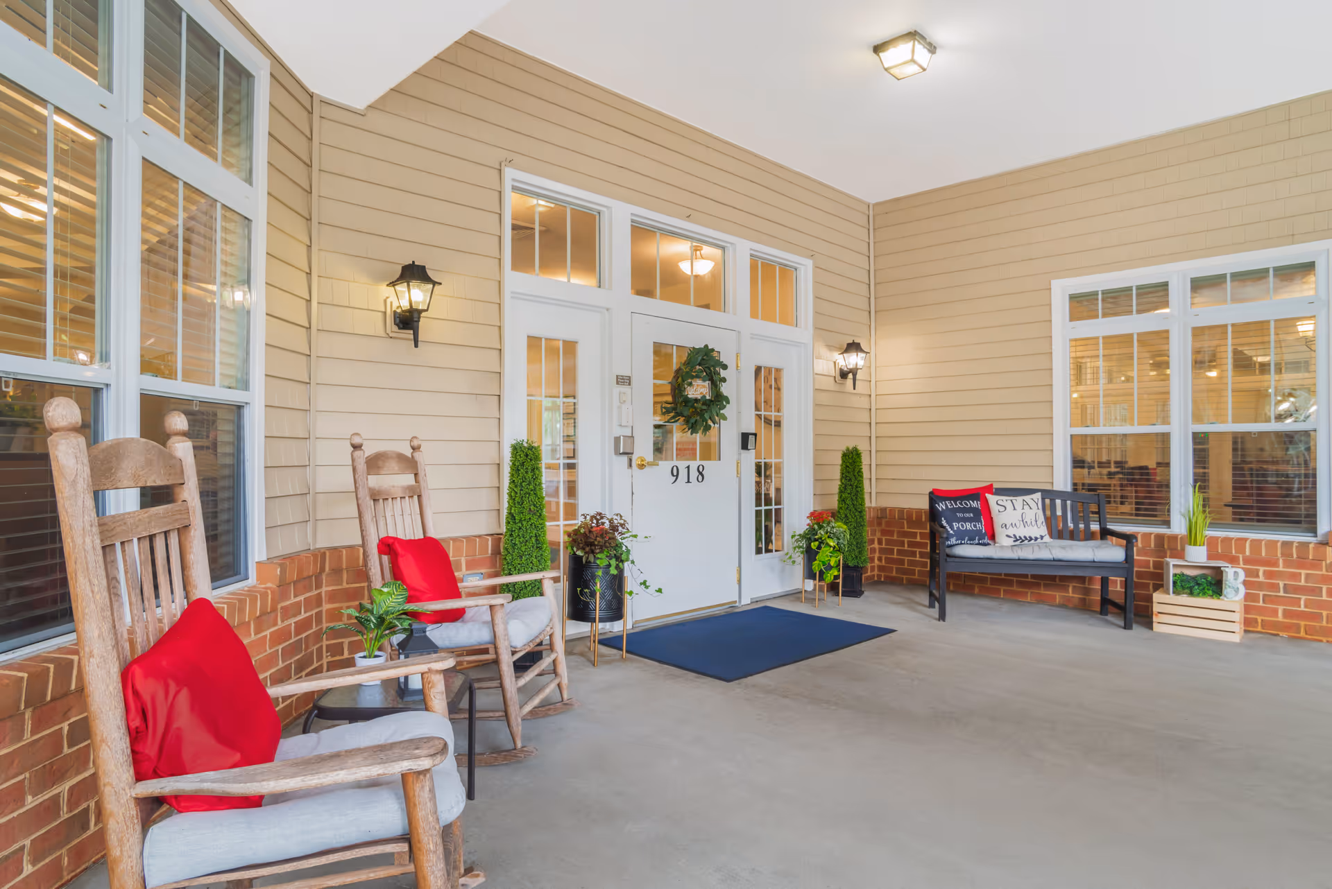 Covered porch area at the entrance of a building with two wooden rocking chairs with red cushions on the left, a black bench with decorative pillows on the right, potted plants, and a white door with the number 918 and a wreath hanging on it.