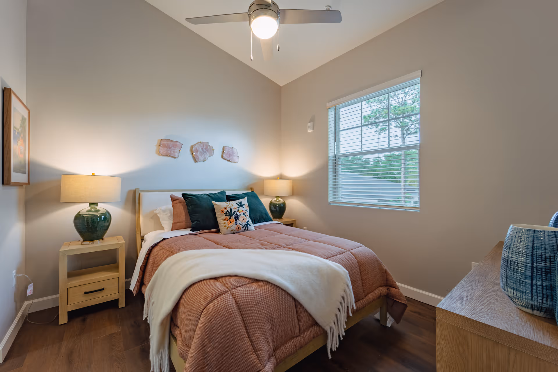 A cozy bedroom with a double bed covered in a rust-colored quilt and white throw blanket. The bed has decorative pillows including a floral one. There are two wooden nightstands on either side of the bed, each with a green ceramic lamp. A window with blinds lets in natural light, and a ceiling fan with a light is on the ceiling. The walls are painted light beige, and there is framed artwork on the left wall and three decorative pieces above the bed.