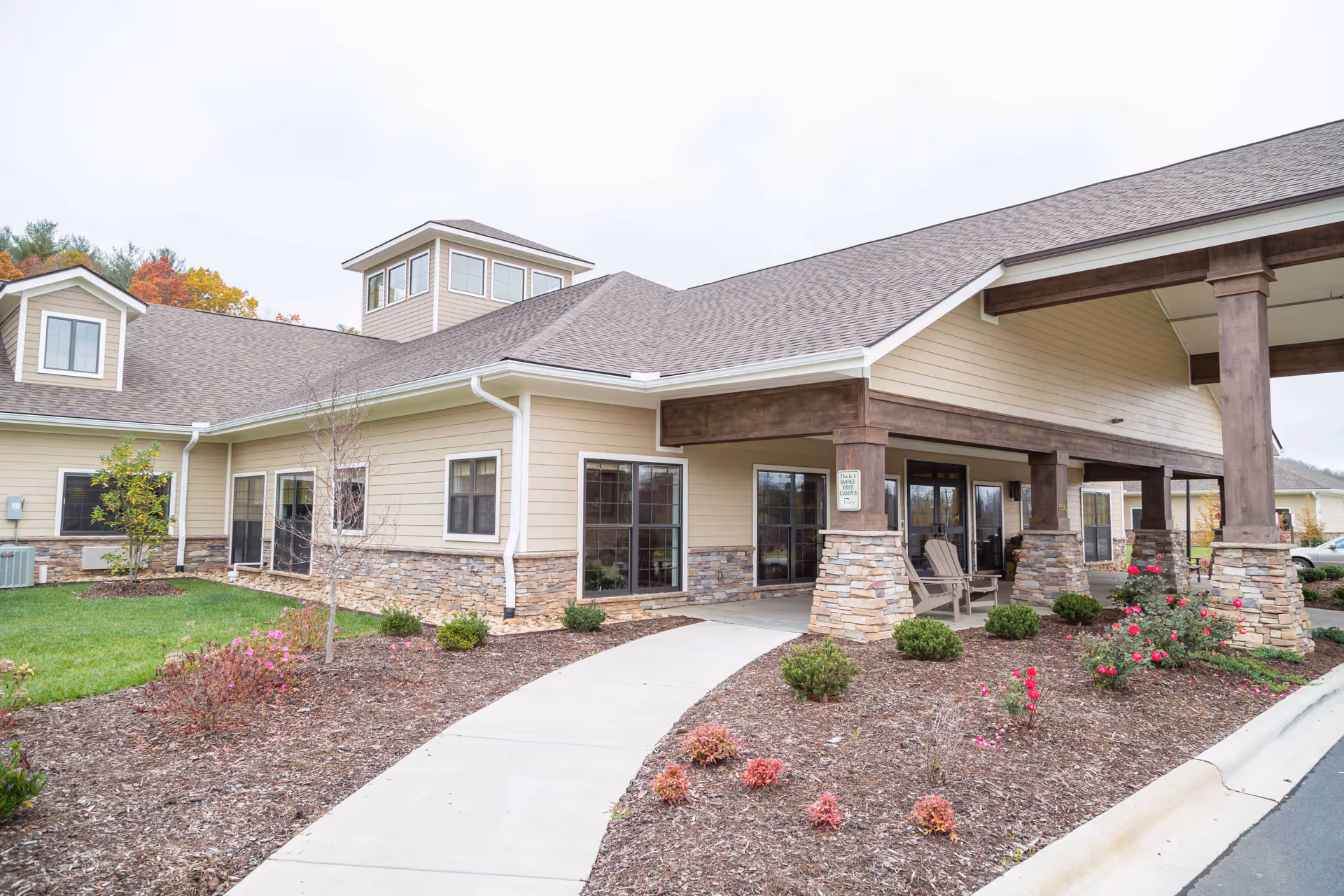 Exterior view of The Lodge at Mills River Health and Rehabilitation building showing a covered entrance with stone pillars, beige siding, multiple windows, landscaped garden beds with shrubs and flowers, and a concrete walkway leading to the entrance.