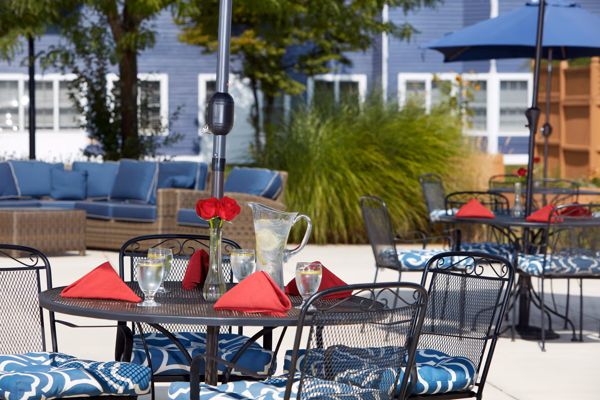 Outdoor patio area with black metal tables and chairs featuring blue and white patterned cushions. Tables are set with red folded napkins, glasses of water with lemon, and a pitcher of water with lemon. In the background, there is a wicker sofa with blue cushions and green plants, with a blue building visible behind.