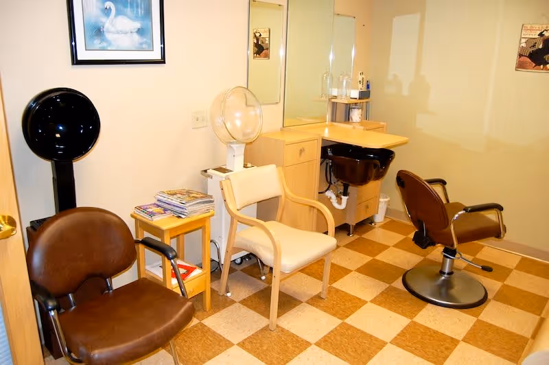 Interior of a small salon or beauty room with two brown salon chairs, a beige chair, a hair dryer, a small table with magazines, a sink with a mirror above it, and light-colored checkered flooring.