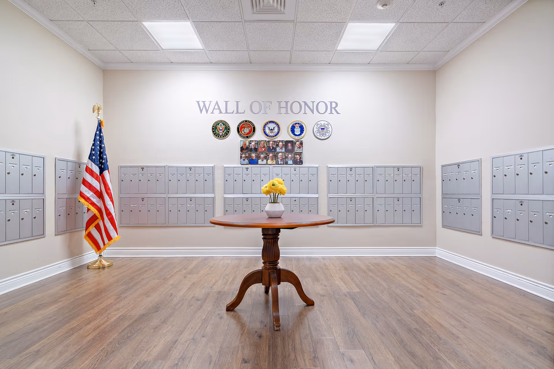 Interior mail room with rows of silver mailboxes, an American flag, a round table with yellow flowers, and a 'Wall of Honor' display on the back wall.