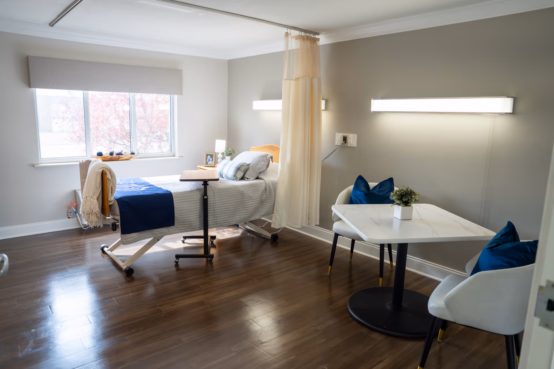 A bright and clean rehabilitation room with a hospital bed near a window, a small overbed table, a privacy curtain, and a seating area with a white table and two white chairs with blue cushions. The room has wooden flooring and neutral-colored walls with modern lighting fixtures.