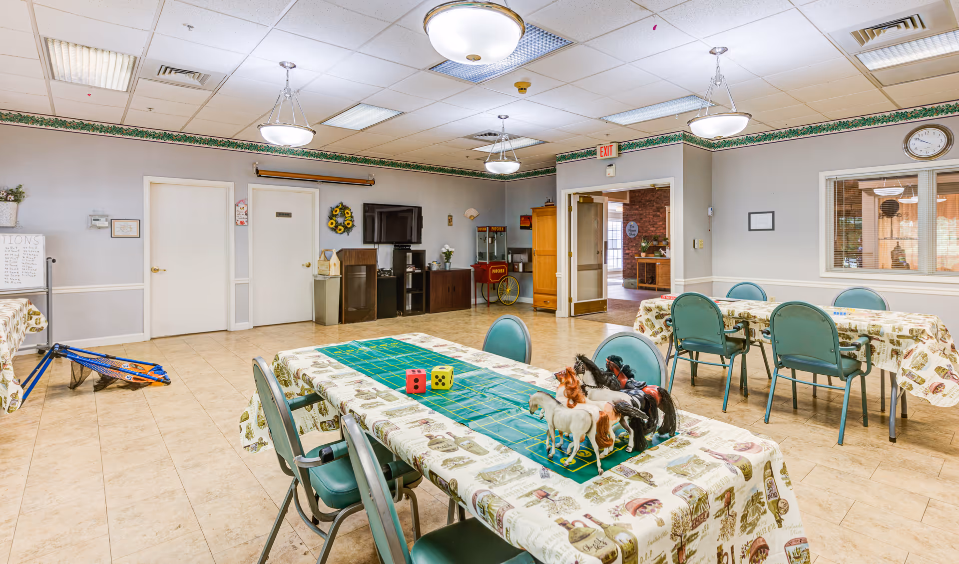 A brightly lit activity room with tables covered in patterned tablecloths. One table has a green game mat, large dice, and several toy horses. The room has tiled floors, light-colored walls with a green border near the ceiling, and several teal chairs around the tables. There is a television mounted on the wall, a popcorn machine, and a doorway leading to another area. Ceiling lights and an exit sign are visible.