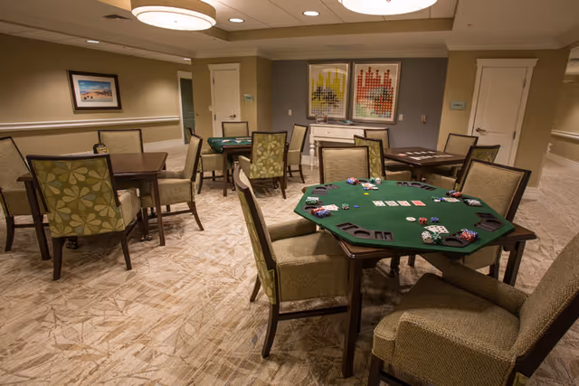 A well-lit room with multiple tables and chairs arranged for card games. One table in the foreground is covered with a green felt poker table top and has playing cards, poker chips, and card holders on it. The room has beige walls, carpeted floors, framed artwork on the walls, and ceiling lights.
