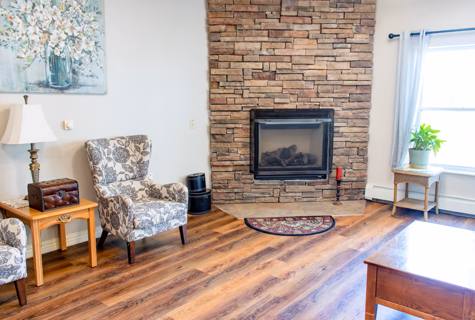 Cozy living room with patterned armchairs, a stone fireplace, hardwood floors, and a side table by a window.