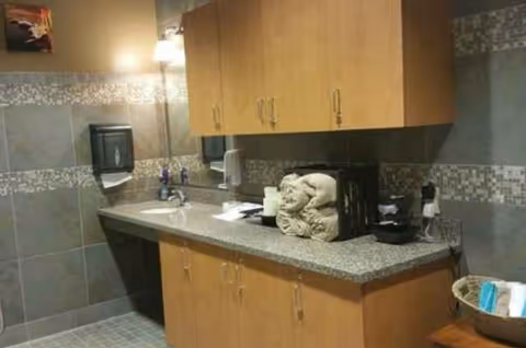 Interior nursing home kitchenette with wooden cabinets, granite counter, sink, coffee maker, and supplies.
