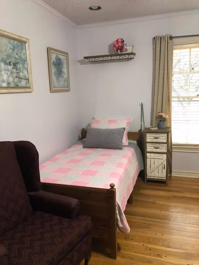 A small bedroom with a single wooden bed covered with a pink and white checkered quilt and two pillows, one pink and one gray. Next to the bed is a rustic white nightstand with three drawers, a small potted plant, and a book on top. A window with beige curtains is on the right wall. On the left side of the bed is a dark upholstered armchair. Two framed floral paintings hang on the left wall, and a small decorative shelf with pink flowers is mounted on the wall above the bed.