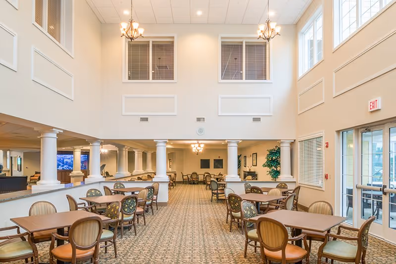 Two-story communal dining room with multiple tables and chairs, chandeliers, columns, and large windows.