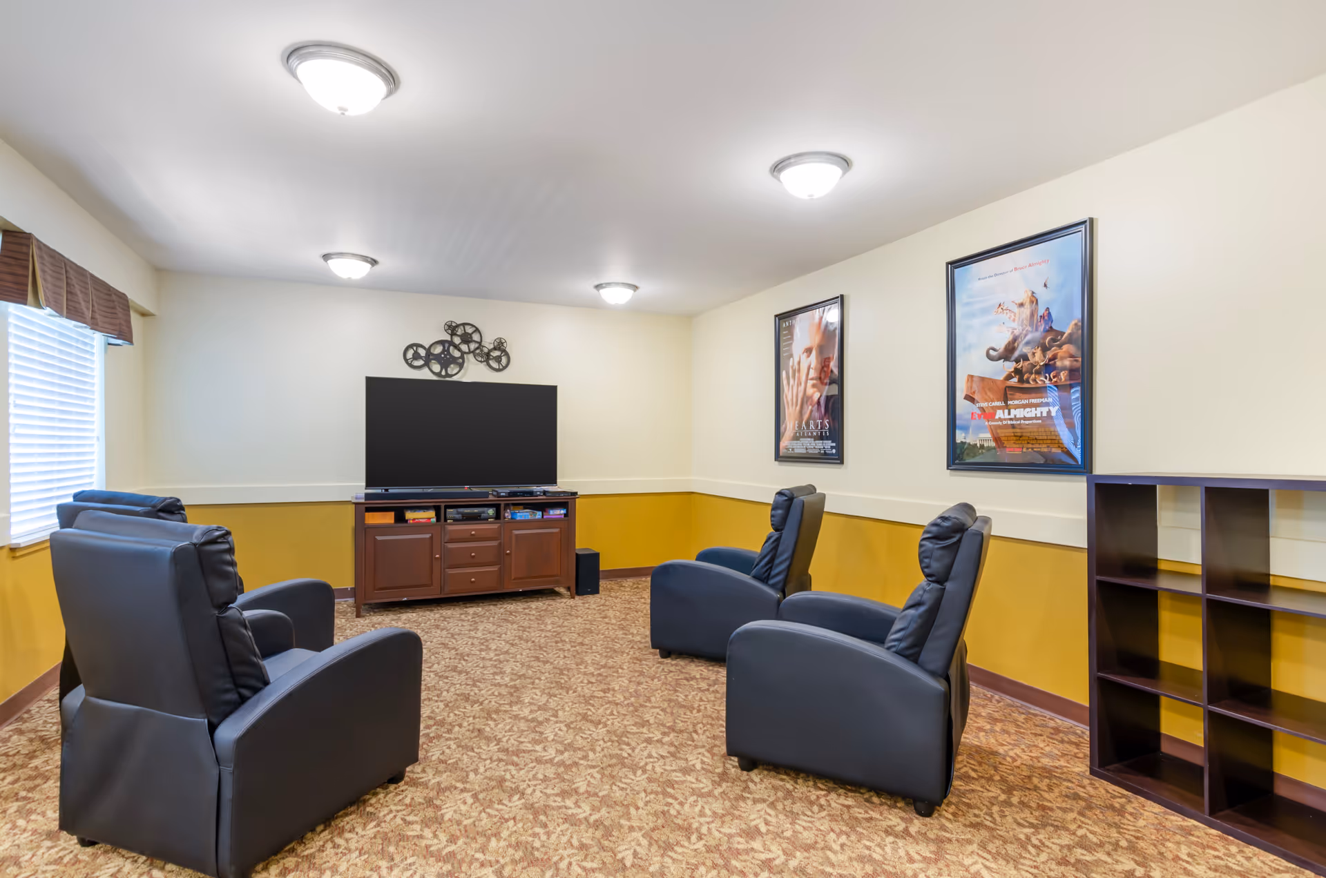 A cozy living room with four black recliner chairs arranged facing a large flat-screen TV on a wooden cabinet. The room has beige walls with a mustard yellow lower half, carpeted floor with a leaf pattern, and three ceiling lights. Two framed movie posters hang on the wall to the right, and a window with blinds and a brown valance is on the left.