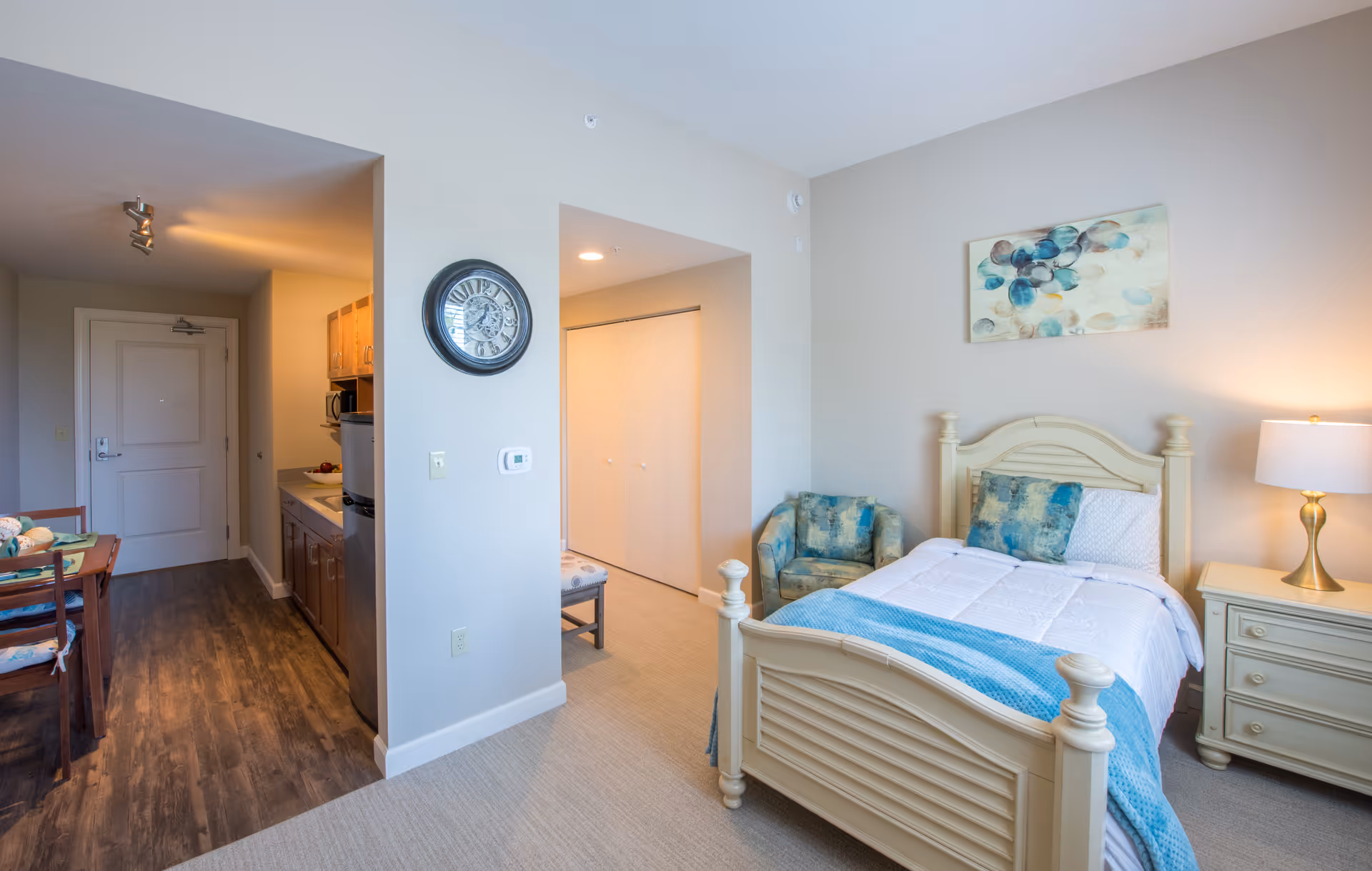 Bright studio-style bedroom with a single bed, nightstand and armchair, with a small kitchenette and dining area visible to the left.
