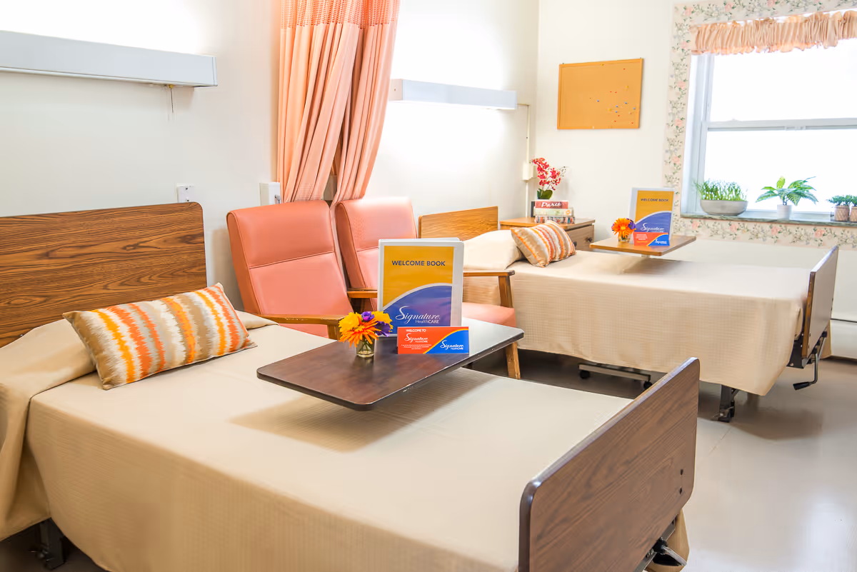 Two hospital-style beds in a bright senior living room with armchairs, overbed tables holding welcome books and small flower arrangements.