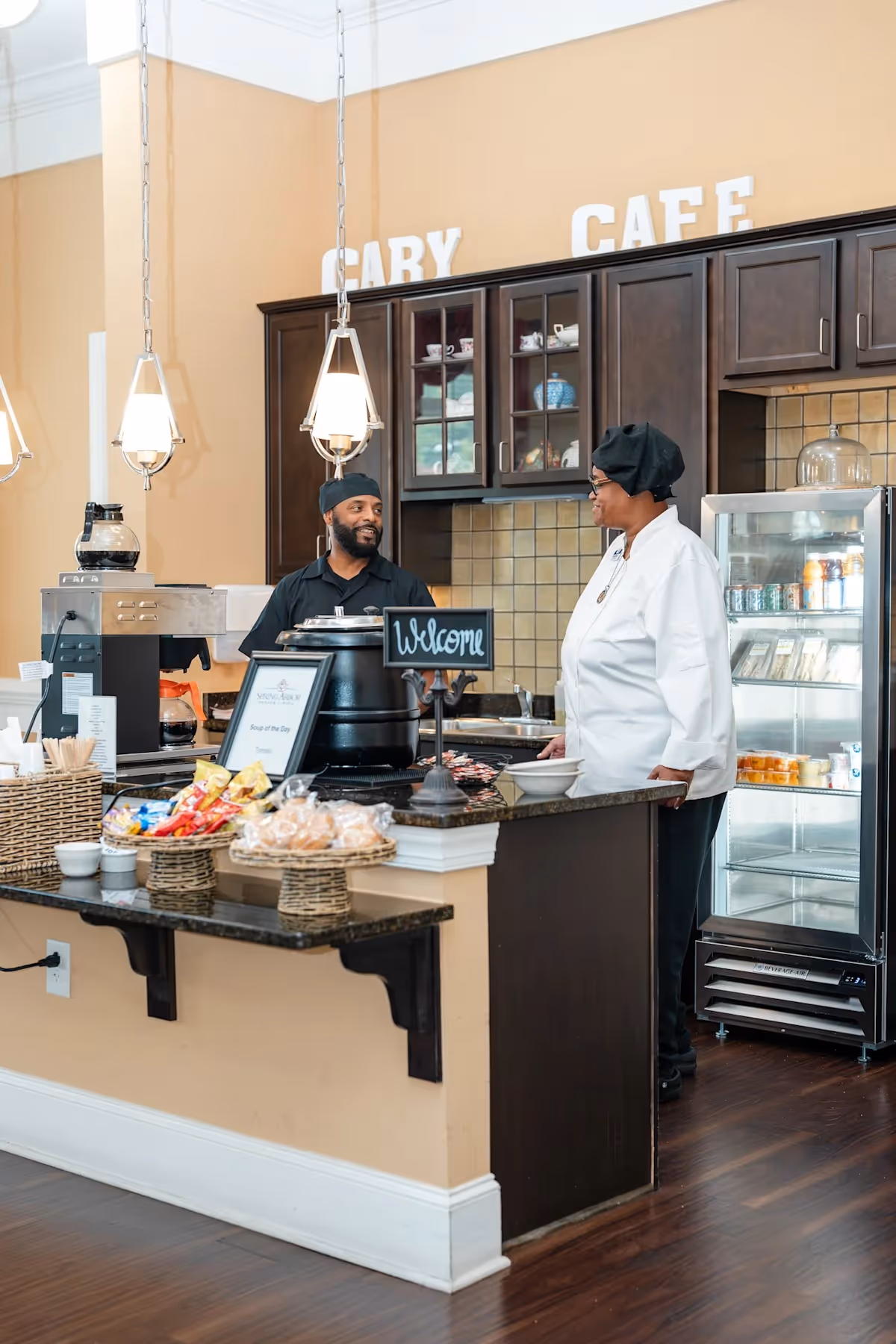Two staff members in a cafe setting at Spring Arbor of Cary. One person is standing behind a counter with snacks and a coffee machine, while the other person, dressed in a white chef's coat and black hat, stands on the customer side of the counter. The background shows dark wooden cabinets, a tiled backsplash, and a refrigerated display case with drinks and food items. Above the cabinets, large white letters spell out 'CARY CAFE'.