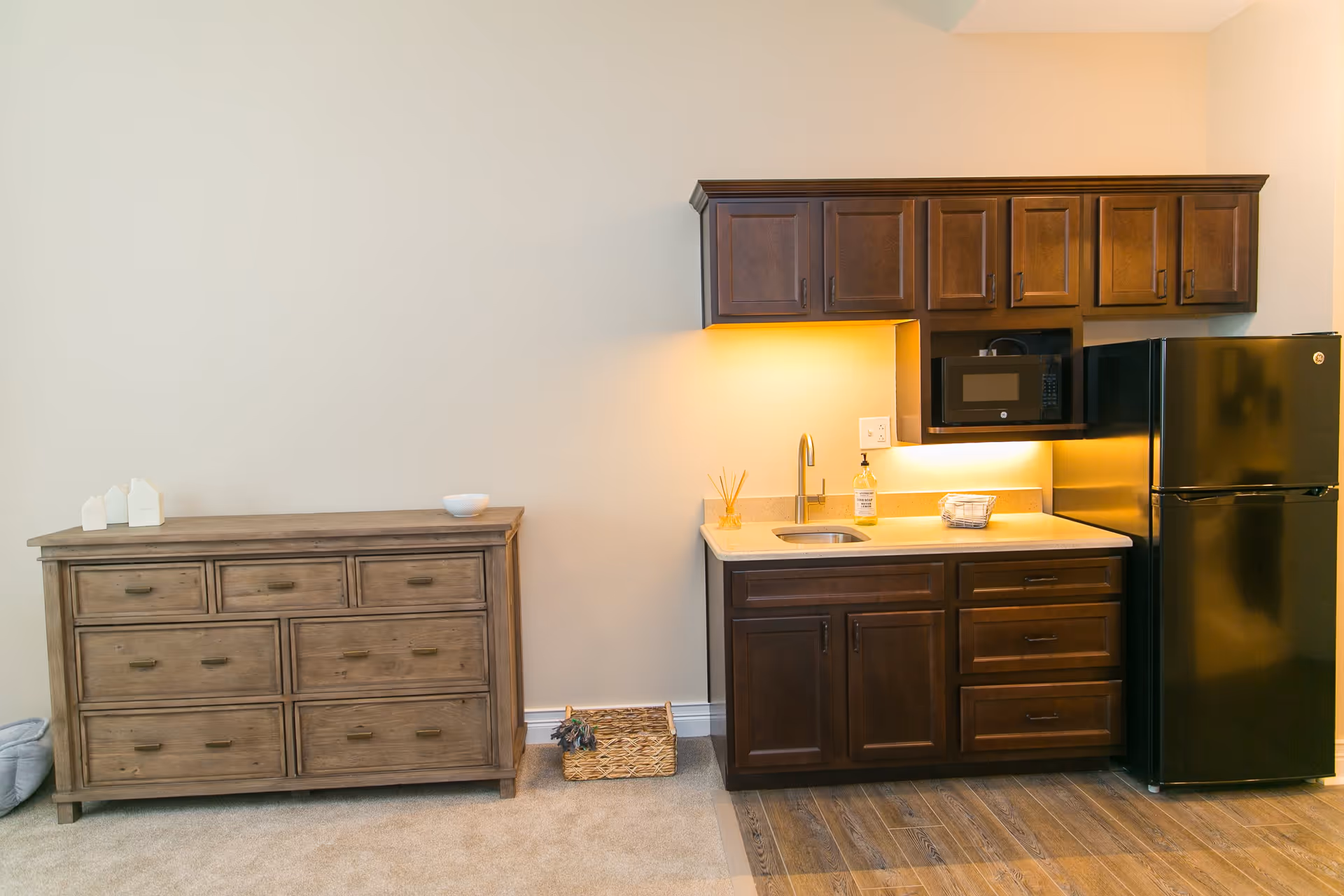 A small kitchenette area with dark wooden cabinets, a black microwave, a black refrigerator, a sink, and under-cabinet lighting. To the left of the kitchenette is a wooden dresser with multiple drawers and a small decorative basket on the floor beside it. The floor transitions from carpet to wood in front of the kitchenette.