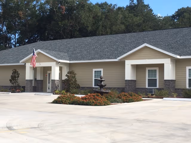 Single-story beige senior living building front with an American flag, fountain, and landscaped driveway.