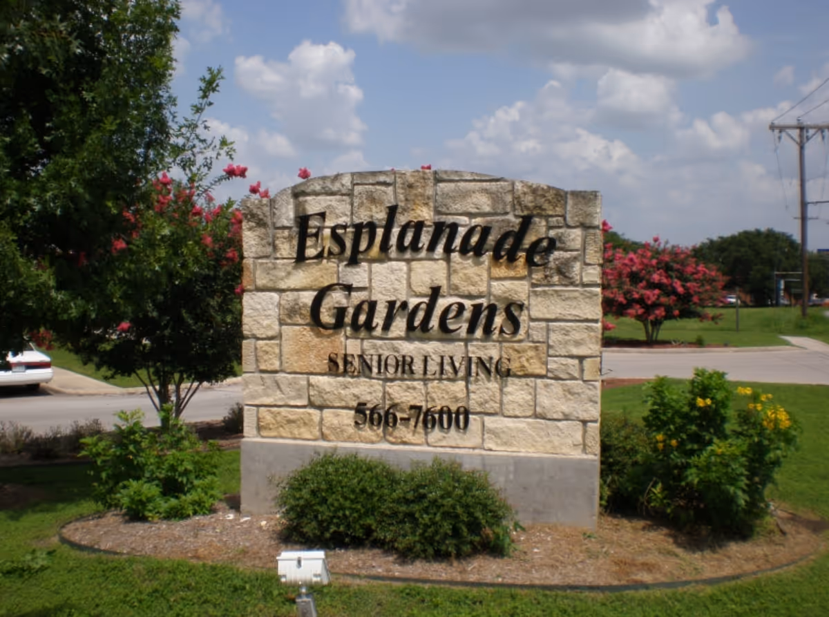 Stone entrance sign reading "Esplanade Gardens Senior Living" surrounded by landscaping and trees.