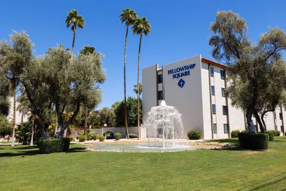 Exterior view of Fellowship Square Tucson building with a water fountain in the foreground, surrounded by green grass, trees, and tall palm trees under a clear blue sky.