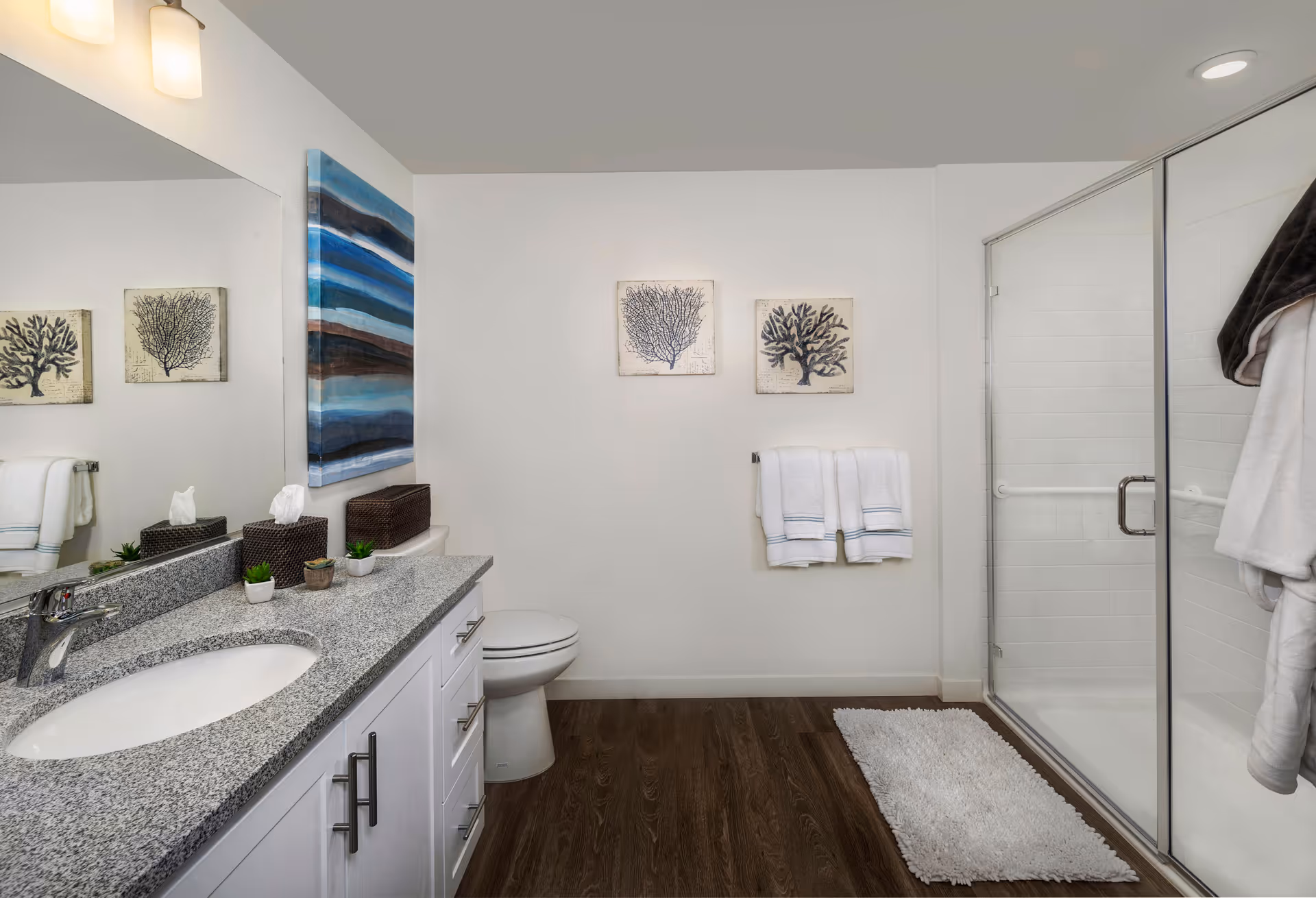 A clean and modern bathroom with a granite countertop sink, white cabinetry, a toilet, and a glass-enclosed shower. The walls are decorated with two framed coral prints and a blue abstract painting. White towels hang on a rack, and a white bath mat is placed on the wooden floor.