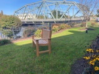 A wooden chair placed on a grassy lawn near a flower bed with yellow flowers. In the background, there is a metal truss bridge over a body of water, with trees and bushes surrounding the area under a clear blue sky.