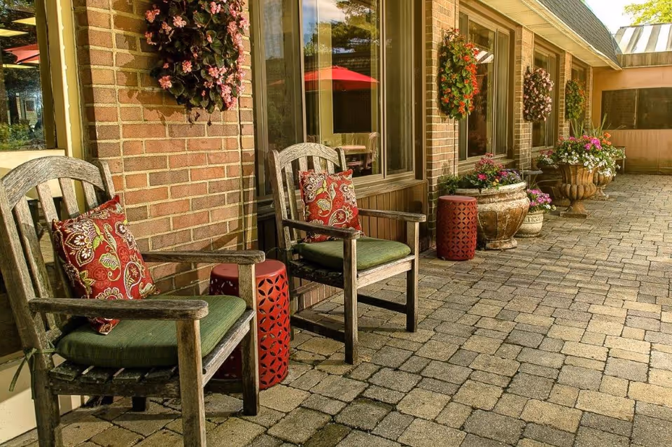 Outdoor patio area with two wooden chairs featuring green cushions and red patterned pillows, a small red decorative side table between them, brick wall with hanging flower baskets, large potted plants, and windows reflecting red umbrellas.