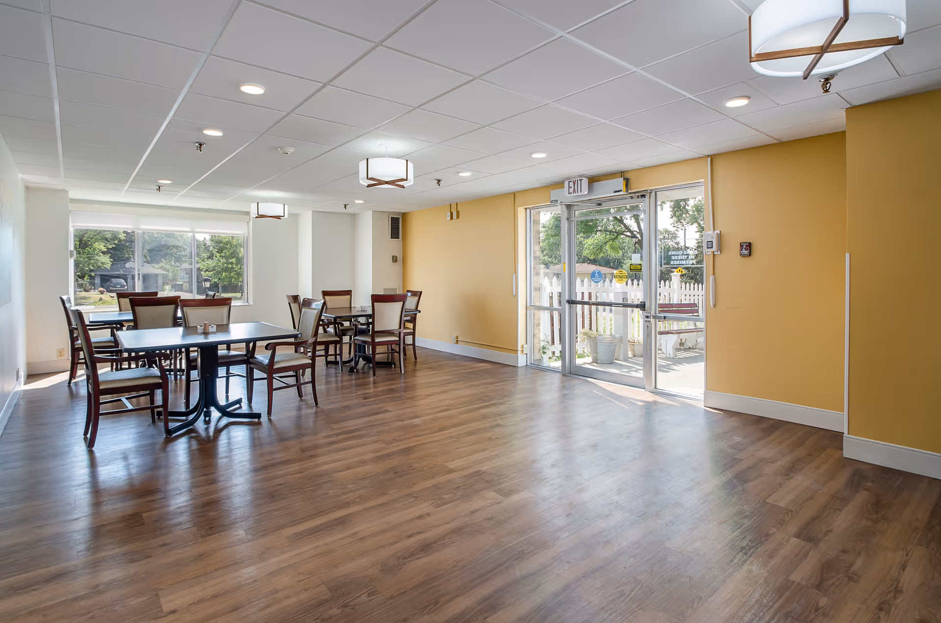 A bright dining area with wooden floors, several tables and chairs, large windows letting in natural light, and glass doors leading outside. The walls are painted yellow and white, and there are ceiling lights providing additional illumination.