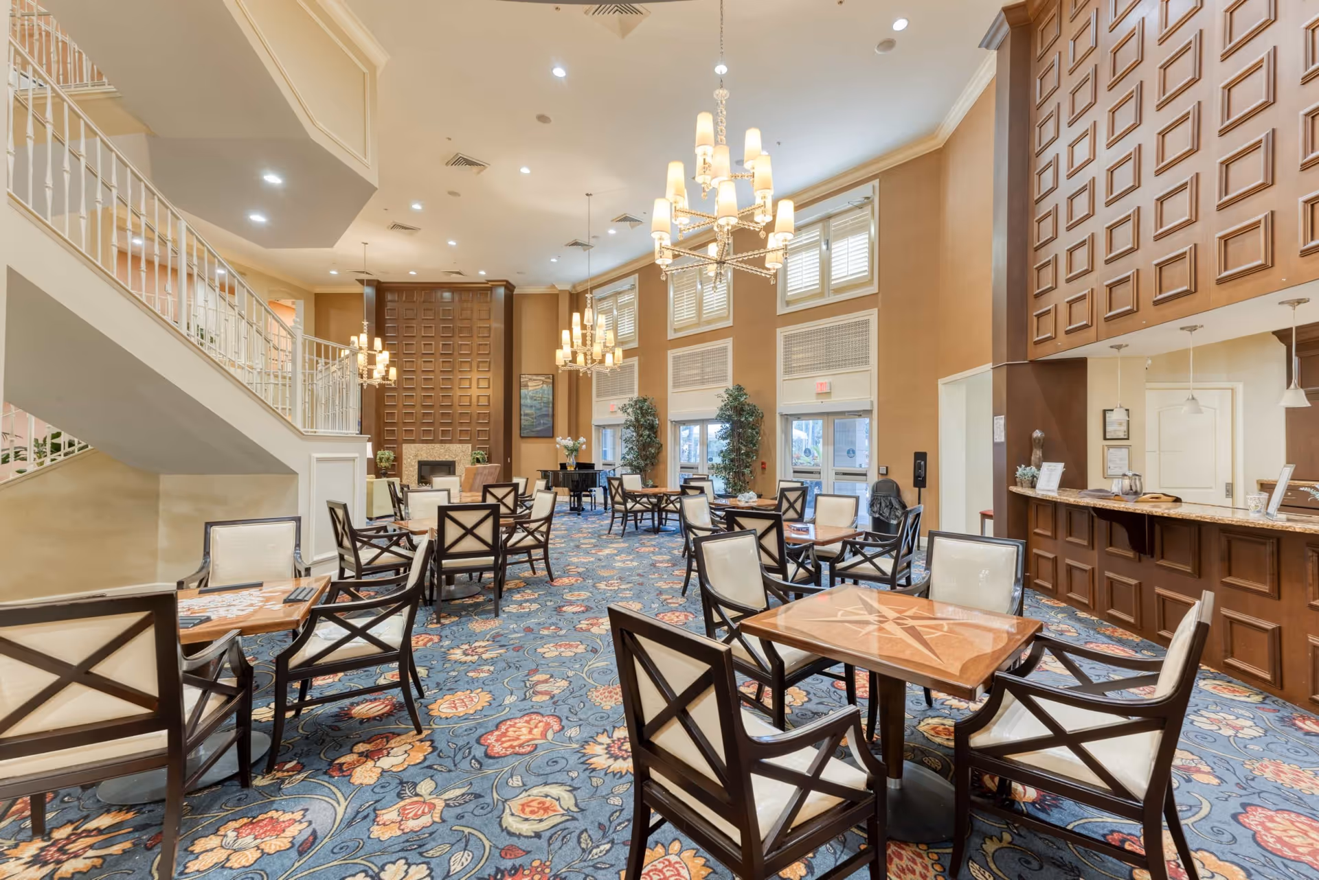 Spacious dining area with multiple wooden tables and chairs arranged on a floral patterned carpet. The room features high ceilings with elegant chandeliers, large windows with shutters, a staircase on the left, and a wooden paneled wall with a fireplace at the far end. There is also a counter area on the right side of the room.