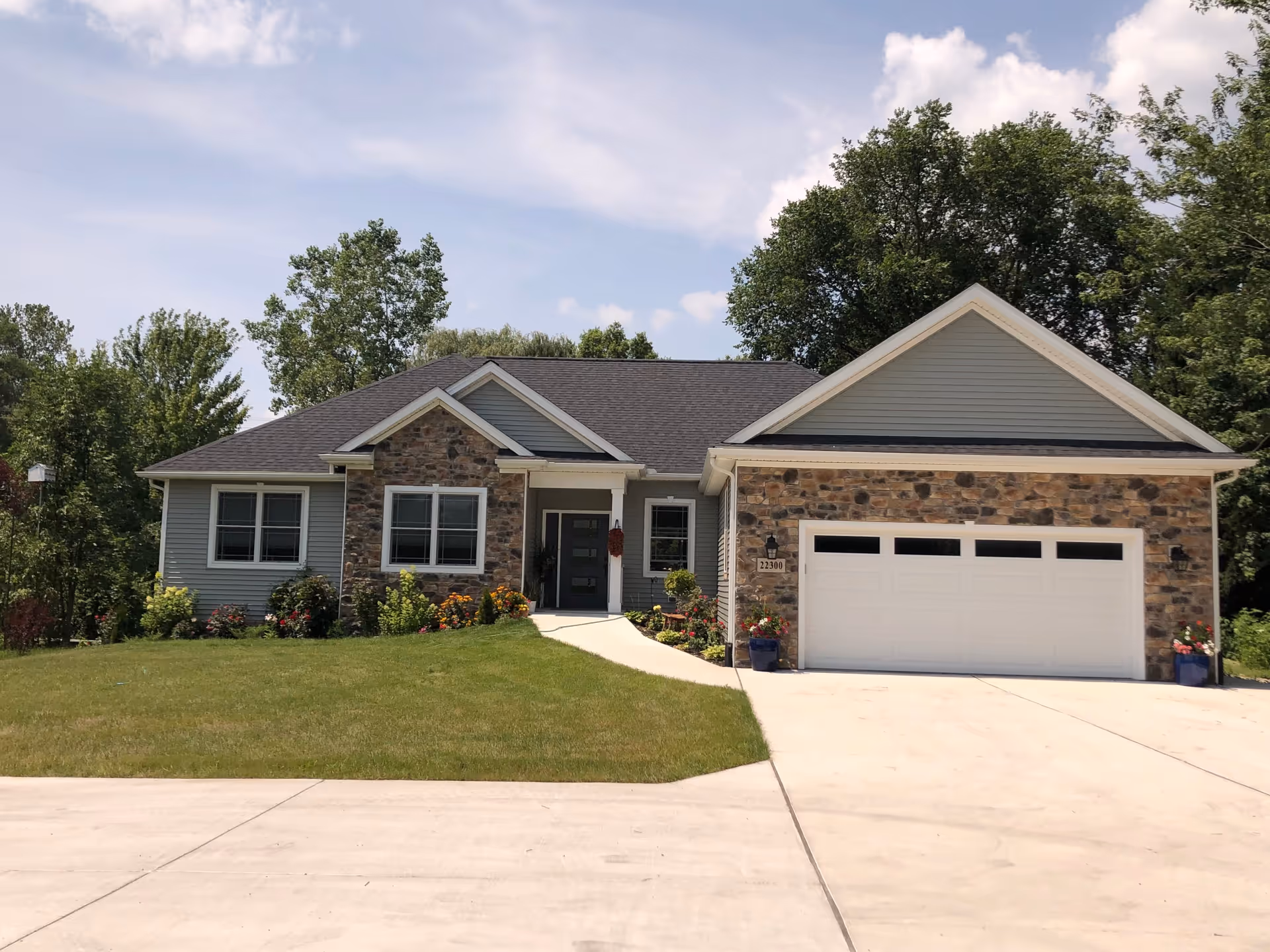 Front exterior view of a single-story house with a stone and gray siding facade, a dark shingled roof, a white garage door, a concrete driveway, and a well-maintained lawn with flowers and shrubs under a partly cloudy sky.