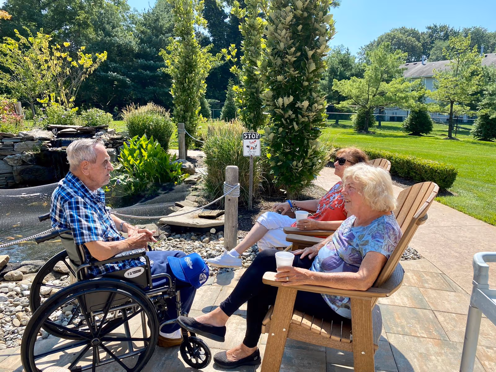 Three elderly individuals enjoying a sunny day outdoors at a senior living facility. One man in a wheelchair is facing two women sitting in wooden Adirondack chairs on a paved patio. They are surrounded by green grass, trees, and landscaping including a small rock garden and a water feature.