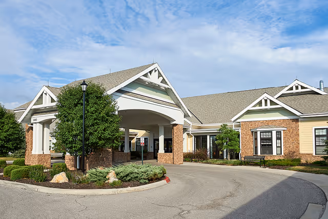 Front entrance of a single-story senior living building with a covered porte-cochère, landscaped island, and clear sky.