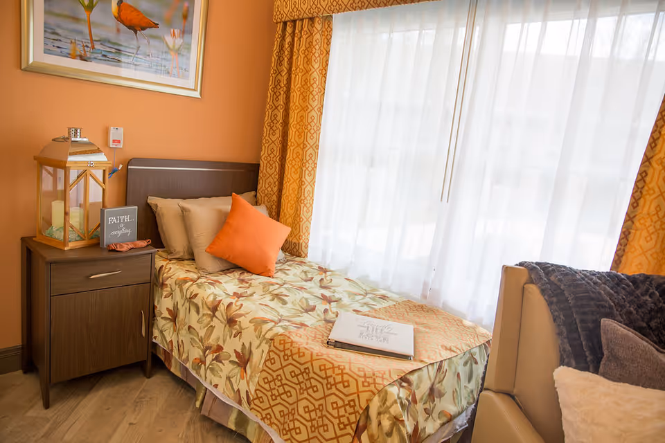 A cozy bedroom with a single bed covered in floral and geometric patterned bedding, an orange throw pillow, and beige pillows. Next to the bed is a wooden nightstand with a large lantern candle holder and a small sign that reads 'FAITH... does everything.' The room has orange walls, a large window with sheer white curtains and patterned orange drapes, and a framed picture of birds on the wall. A beige armchair with a dark gray throw blanket is partially visible.