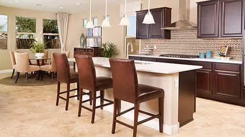 Open-plan kitchen with a large white island and three brown bar stools, dark wood cabinets, and a dining area by windows.