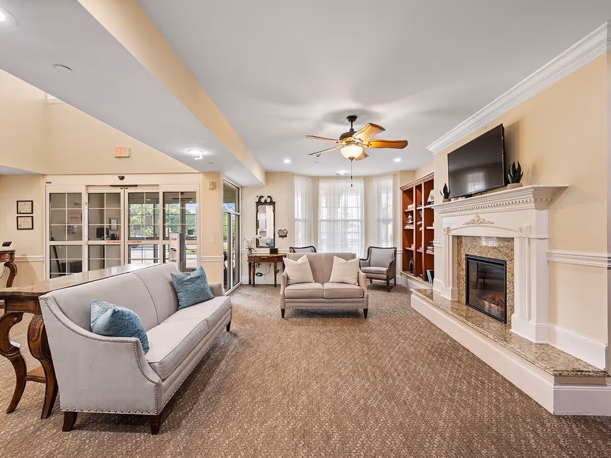 A bright and inviting living room area in a senior living facility featuring beige walls, a ceiling fan with lights, a fireplace with a mounted flat-screen TV above it, a built-in wooden bookshelf, and comfortable seating including a gray sofa with blue pillows, a loveseat with white pillows, and an armchair. Large windows with white blinds allow natural light to fill the room. The entrance with glass doors is visible in the background.