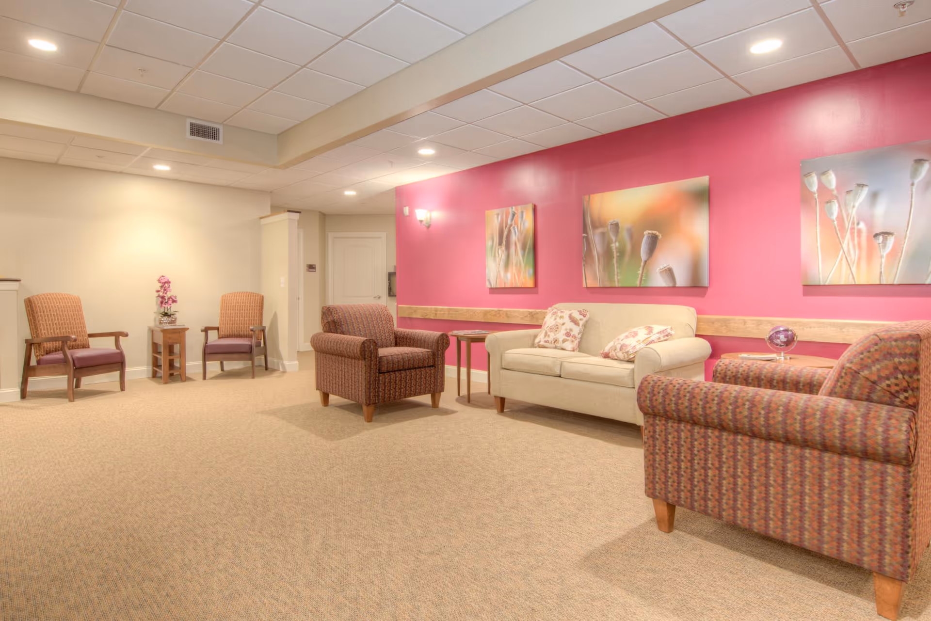A cozy seating area in a senior living facility with a beige couch adorned with floral pillows, two patterned armchairs, and two wooden chairs with purple cushions. The walls are painted beige and bright pink, with three floral-themed paintings hanging on the pink wall. The room is well-lit with recessed ceiling lights and a wall sconce, and there is a small wooden side table with a purple flower arrangement between the two wooden chairs.
