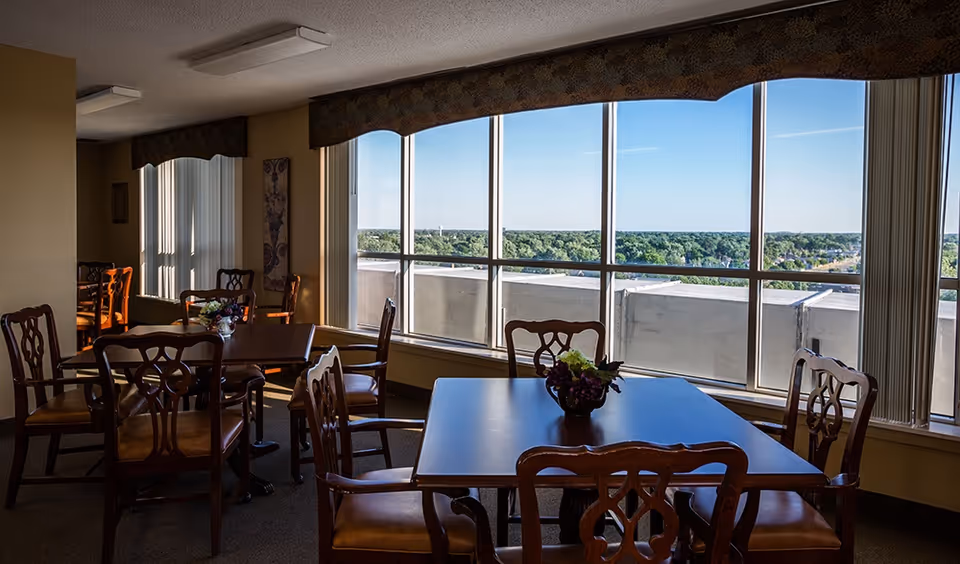 Dining room with wooden tables and chairs, floral centerpieces, and large windows overlooking treetops.