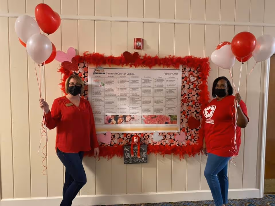 Two women wearing red tops and black face masks stand on either side of a decorated bulletin board with a February 2021 calendar for Savannah Court of Camilla. Both women hold red and white balloons. The bulletin board is decorated with red feathers and heart-themed paper, and there is a small welcome sign below it.